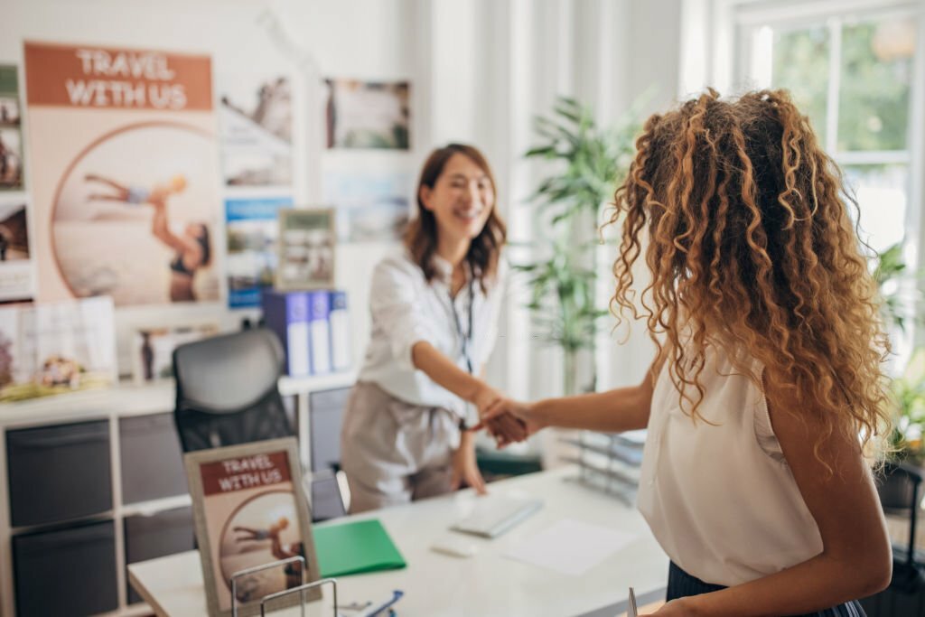 Two people, a beautiful woman arriving in a modern travel agency and shaking hands with a travel agent.